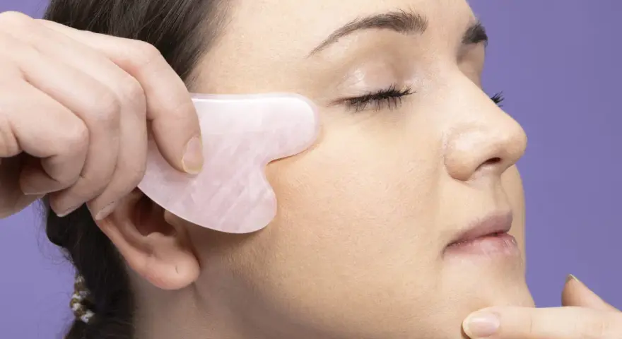 Eine Hand nutzt ein Gua Sha Stein für eine Gesichtsmassage. Das Werkzeug ist herzförmig und aus rosafarbenem Stein.