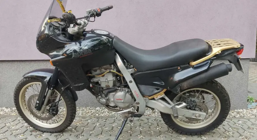 Czarny motocykl Aprilia Pegaso 650 z złotym bagażnikiem i lusterkami, zaparkowany na kostce brukowej.