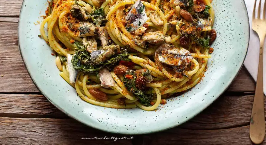 Un piatto di pasta con le sarde, condita con erbe aromatiche, pinoli e pangrattato tostato, servita su un piatto azzurro.