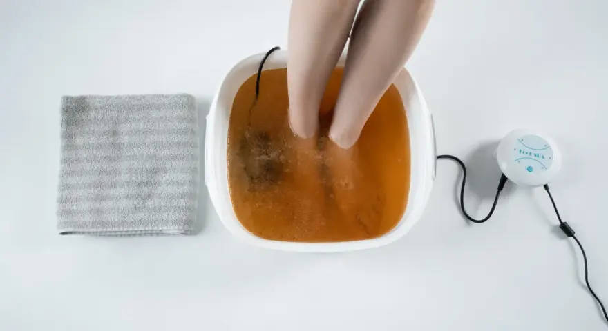 A person's feet are soaking in a foot spa with a brown solution, suggesting a method for how to remove toxins from feet naturally. A towel is nearby.