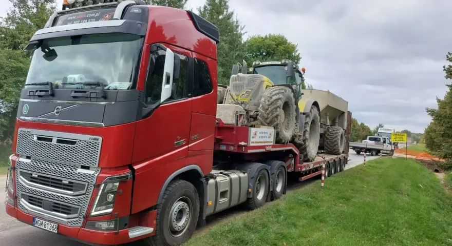 Czerwony ciągnik siodłowy przewozi koparkę na naczepie. To transport ponadgabarytowy, który wymaga specjalnych zezwoleń i przygotowania.