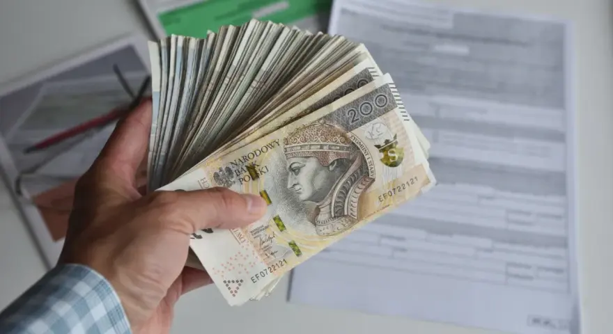 Dłoń trzyma plik banknotów 200 zł. W tle dokumenty, być może dotyczące tego, ile godzin to 0.5 etatu.