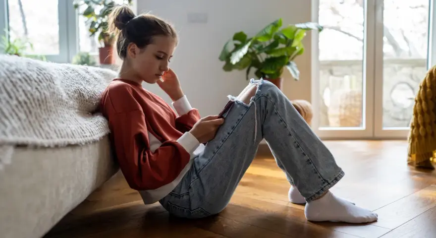 En ung tjej sitter på golvet, försjunken i sin telefon. Hennes hållning och ansiktsuttryck kan tyda på depression hos barn.