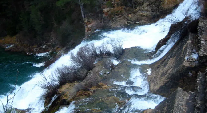 El salto de Poveda, una cascada de agua blanca y espumosa que desciende por rocas y vegetación, desemboca en un río de aguas turquesas.