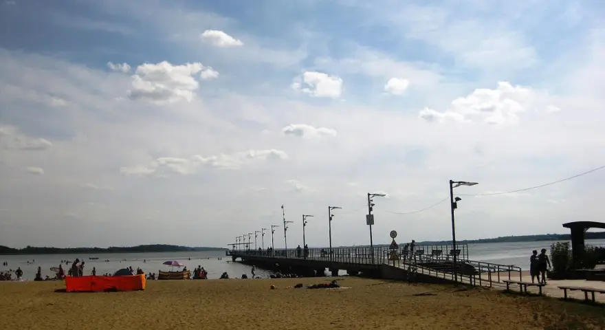 Plaża nad jeziorem Miedwie, z molo i spacerującymi ludźmi. Słoneczny dzień, chmury na niebie.
