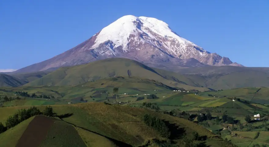 Pokryty śniegiem wulkan Chimborazo, jeden z najwyższych gór świata, góruje nad zielonymi, pofałdowanymi wzgórzami z polami uprawnymi.