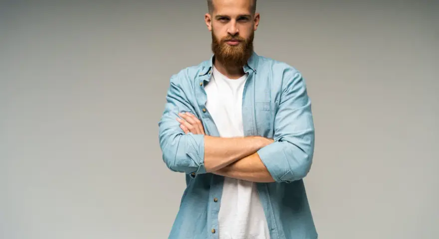 Uomo con barba e capelli biondi, camicia azzurra aperta su t-shirt bianca, braccia conserte. Look trasandato ma curato.
