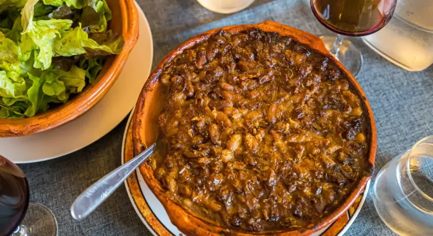 Cassoulet de Castelnaudary burbujeante en cazuela de barro, acompañado de ensalada verde y vino tinto.