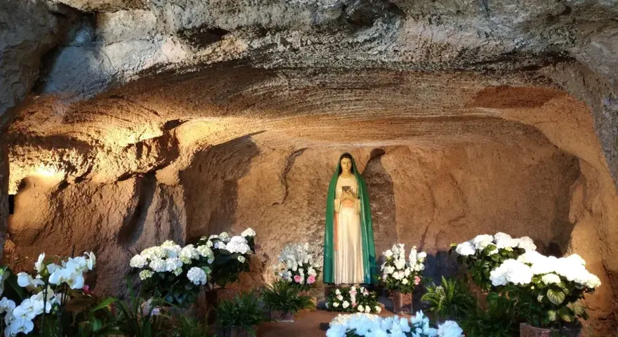 A statue of Mary stands in a grotto, surrounded by lush white flowers. The cave walls are illuminated, creating a serene atmosphere, reminiscent of the three fountains of grace.