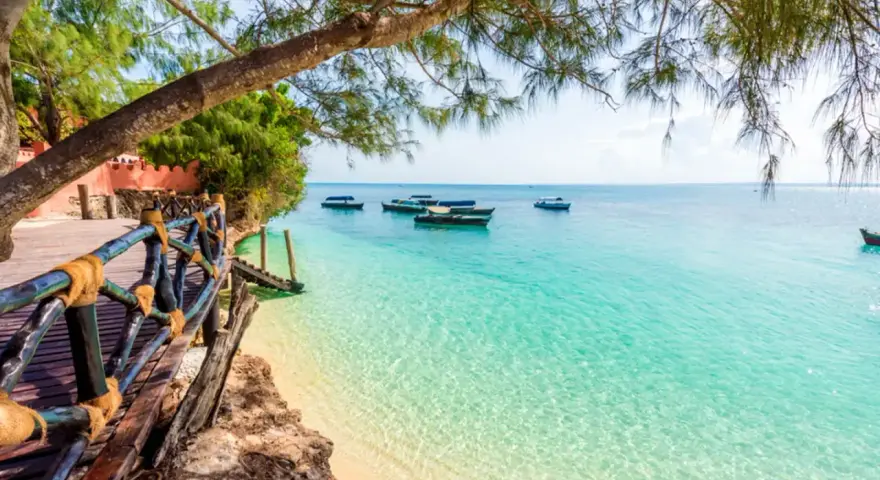 Drewniana kładka prowadzi do krystalicznie czystej wody na plaży. Łodzie kołyszą się na turkusowym morzu. Rajskie wakacje na Zanzibarze.