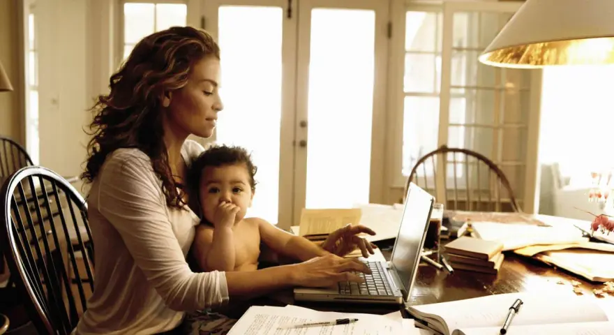 Una mamma lavoratrice con il suo bambino in grembo, mentre digita al computer tra libri e fogli.