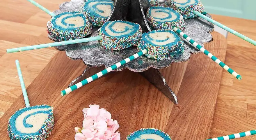 Blå och vita cake pops med strössel på ett silverfärgat fat. Vissa ligger på ett träunderlag med rosa blommor.