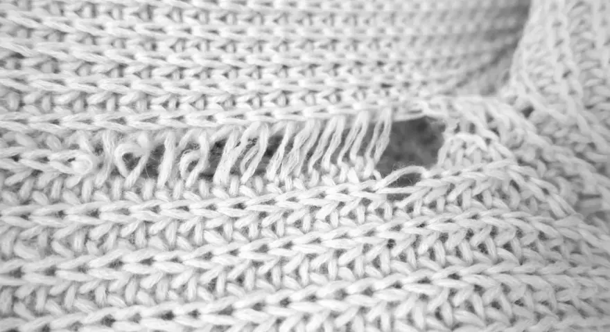Nahaufnahme eines weißen Strickpullovers mit einem Loch und ausgefransten Fäden. Was tun, wenn die Wolle eingelaufen ist?