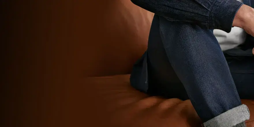Uomo con jeans scuri seduto su divano in pelle. Scopri come lavare jeans a mano per mantenerli perfetti.