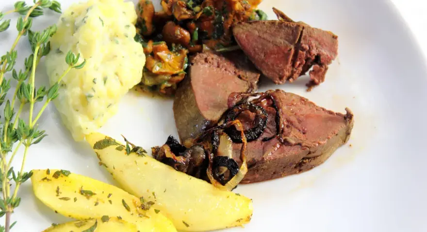Rehleber zubereiten: zarte Scheiben Rehleber mit gebratenen Zwiebeln, Kartoffelpüree, gebratenen Kartoffelspalten und frischen Kräutern auf einem weißen Teller.