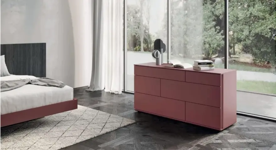 Cassettiera camera da letto moderna color bordeaux, con sei cassetti, accanto a un letto e una grande finestra.