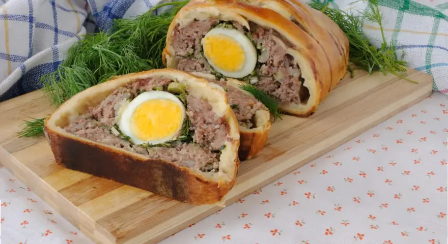 Une tranche de pâté de Pâques, garnie d'un œuf dur, est présentée sur une planche en bois, avec de l'aneth frais.