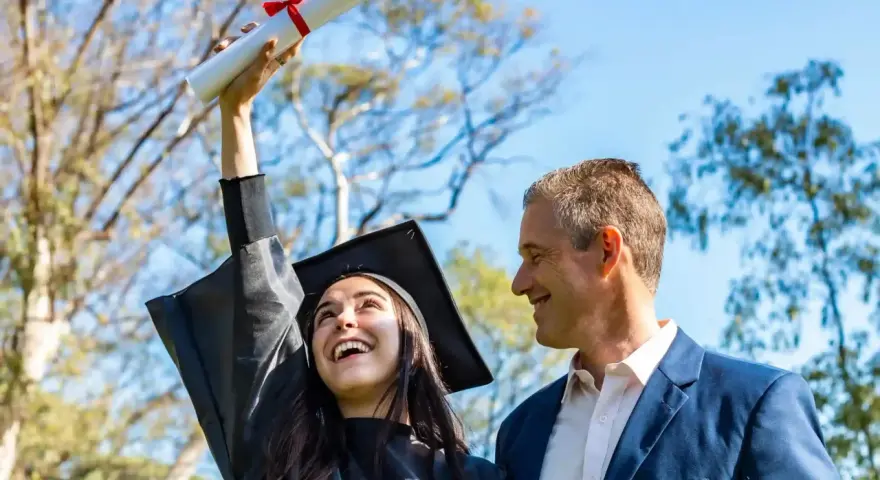 Radosna absolwentka w czapce i todze trzyma dyplom, a obok niej uśmiechnięty mężczyzna. To piękne przykładowe gratulacje z okazji ukończenia studiów.
