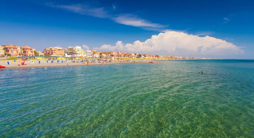 Słoneczna plaża w Rzymie, pełna kolorowych parasoli i ludzi kąpiących się w krystalicznie czystej wodzie.