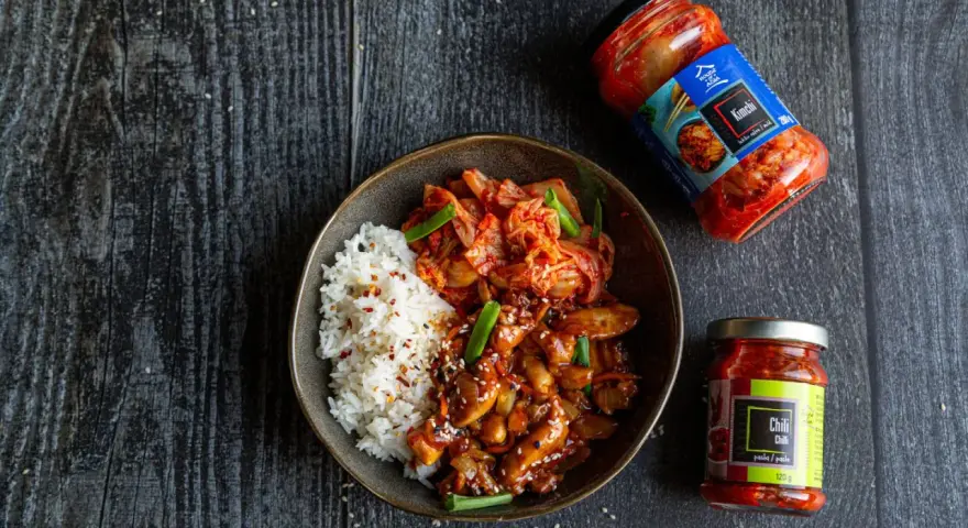 Miseczka ryżu z kurczakiem w sosie i kimchi, obok słoiczki z kimchi i pastą chili.