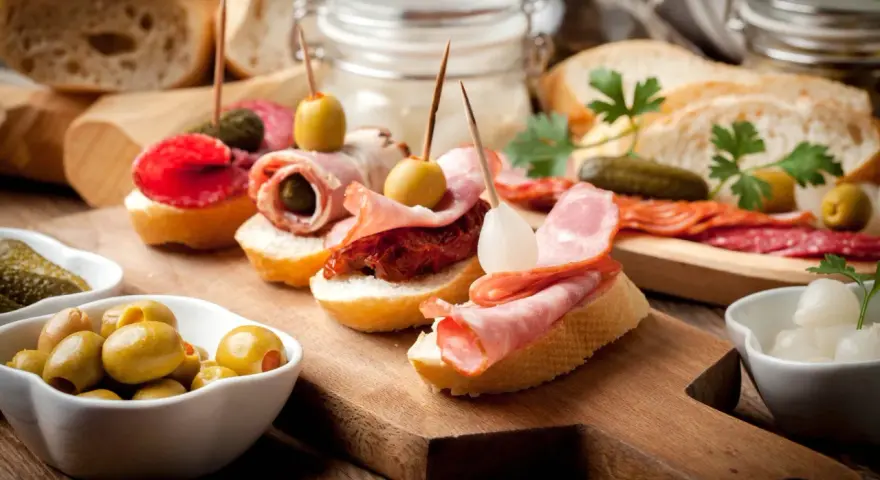 Pyszne tapas na drewnianej desce: pieczywo z wędliną, oliwkami, ogórkiem i suszonymi pomidorami.