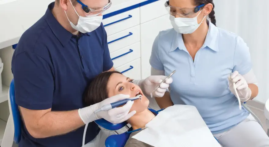 Dentysta i asystentka w okularach i maseczkach pracują przy pacjentce. Asystentka trzyma narzędzie, a dentysta używa **vector paro** do zabiegu.