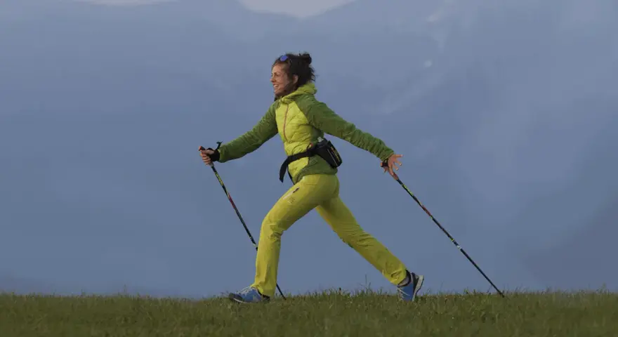 Donna sorridente pratica nordic walking su un prato. Informarsi sulle controindicazioni prima di iniziare.