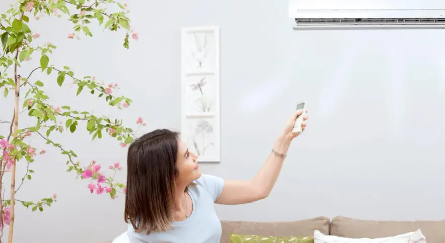 Mujer ajusta el fan speed del aire acondicionado con el control remoto, rodeada de una planta con flores rosas.