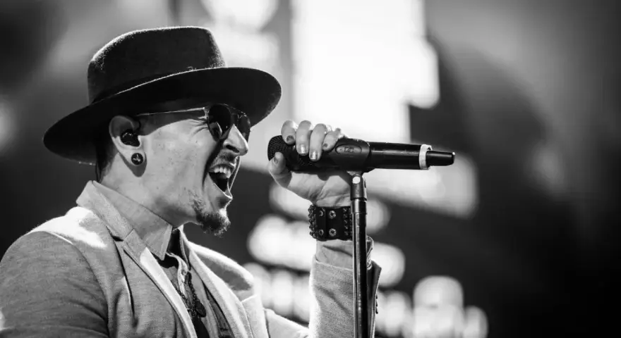 Wokalista Linkin Park, Chester Bennington, w kapeluszu i okularach przeciwsłonecznych, śpiewa do mikrofonu z pasją.