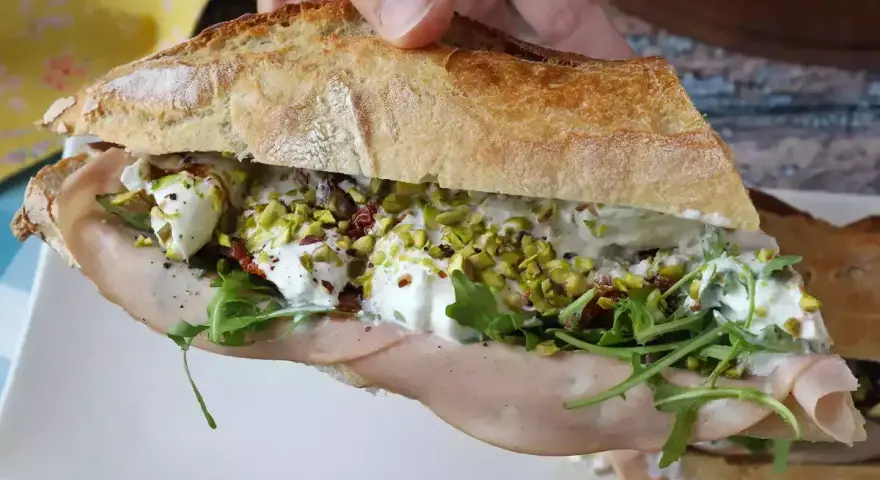 Bocadillo de pan rústico relleno de mortadela, burrata cremosa, rúcula, pistachos y tomates secos.