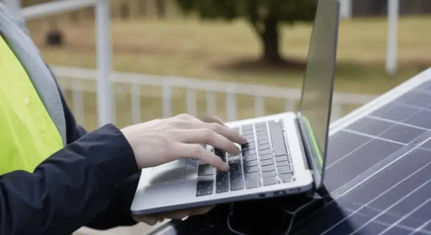 Praca nad projektem fotowoltaiki: ręce na klawiaturze laptopa, obok panel słoneczny.
