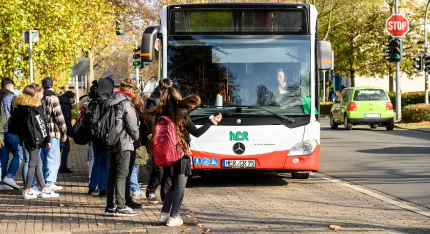 Ein Schulbus mit Warnblinker hält an einer Haltestelle. Schüler warten auf dem Gehweg, um einzusteigen.
