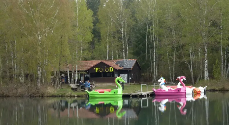Bunte Tretboote in Form von Schwänen und Einhörnern liegen am Ufer des Bruchsee Duingen. Ein Gebäude mit der Aufschrift "FOOD" und Solaranlagen steht im Hintergrund.