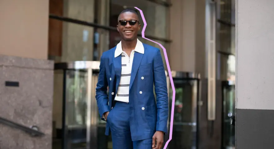 Hombre sonriente con traje azul y camiseta blanca a rayas, gafas de sol, posando con la mano en el bolsillo.