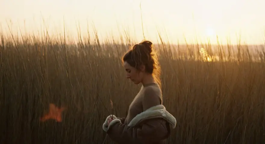 Une jeune femme, le regard baissé, se tient dans les hautes herbes au crépuscule. La lumière dorée du soleil couchant illumine ses cheveux et le paysage.