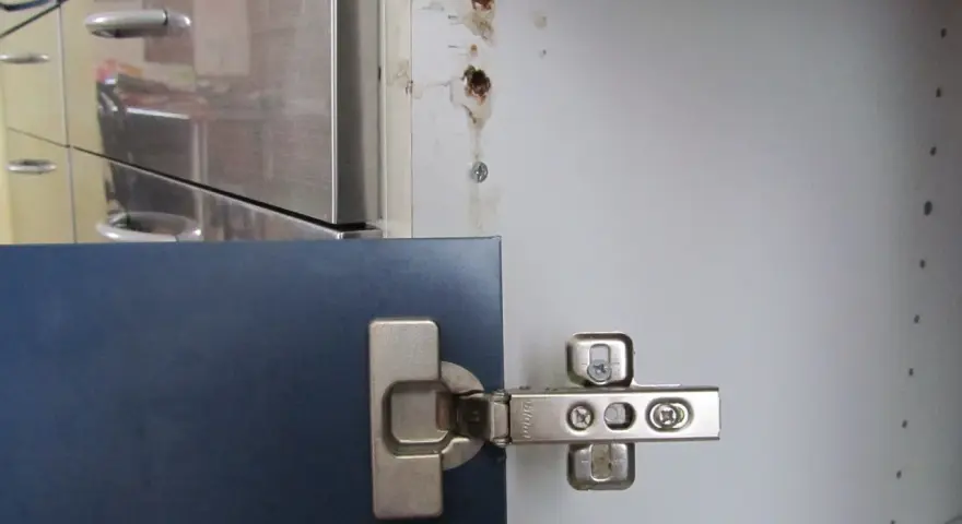 Cerniera di un mobile blu fissata con viti. Vicino, un foro nel legno indica come avvitare una vite che gira a vuoto nel legno.