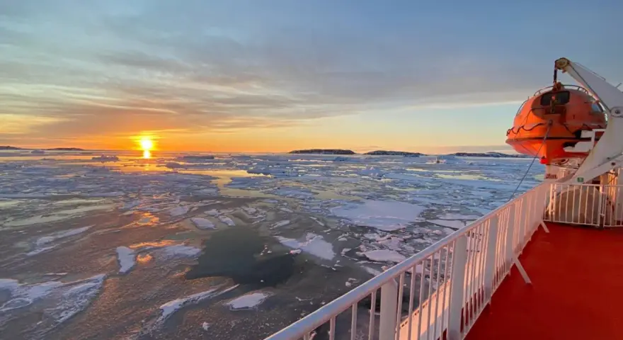 A **Greenland ferry** navigates icy waters at sunset. An orange lifeboat hangs ready on the ship's deck.