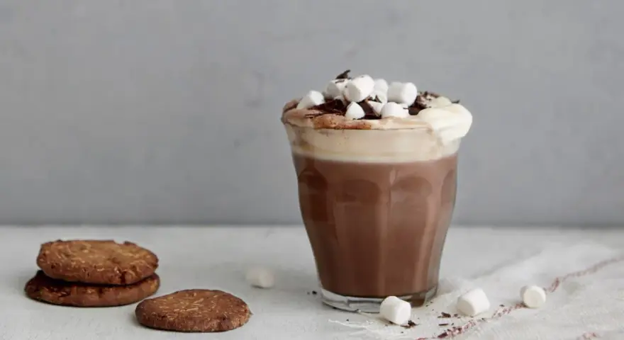 En härlig varm choklad med choklad, toppad med marshmallows och chokladspån. Bredvid står några chokladkakor.
