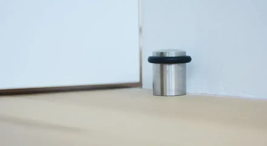 Metalowy odbojnik z czarną gumką przy ścianie, chroniący drzwi przed uderzeniem.