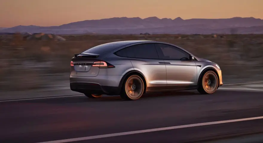 Srebrny Tesla X pędzi przez pustynny krajobraz o zachodzie słońca. Zastanawiasz się nad jego ceną?