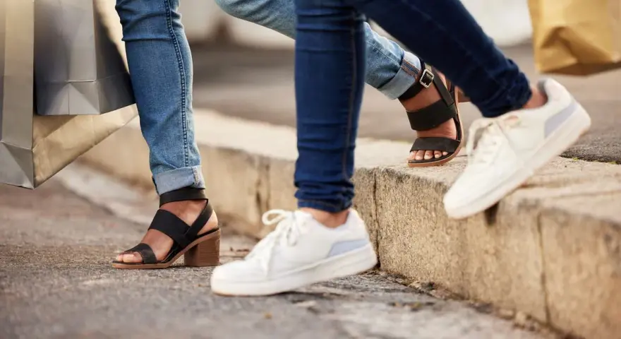 Dwie osoby w jeansach i modnych butach, z torbami na zakupy w rękach, idą chodnikiem. Jedna ma czarne sandały na obcasie, druga białe sneakersy.