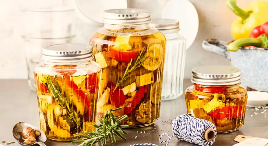 Drei Gläser mit italienischem Fingerfood: eingelegtes Gemüse wie Paprika und Zucchini mit Kräutern in Öl.
