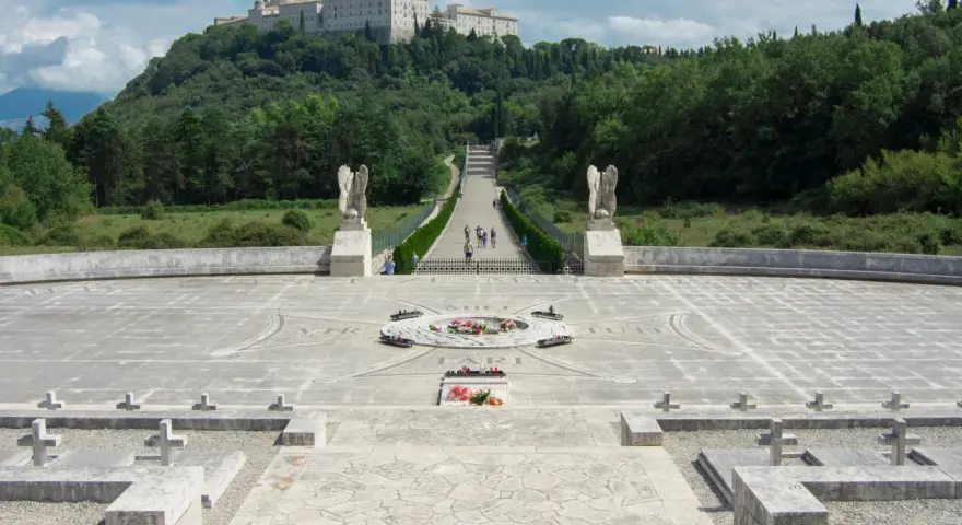 Cmentarz żołnierzy na wzgórzu, z widocznym klasztorem na szczycie. Droga prowadzi do niego przez monumentalny plac z krzyżami i pomnikiem. To miejsce pamięci o bitwie pod **Cassino - trzeci / rzeka**.