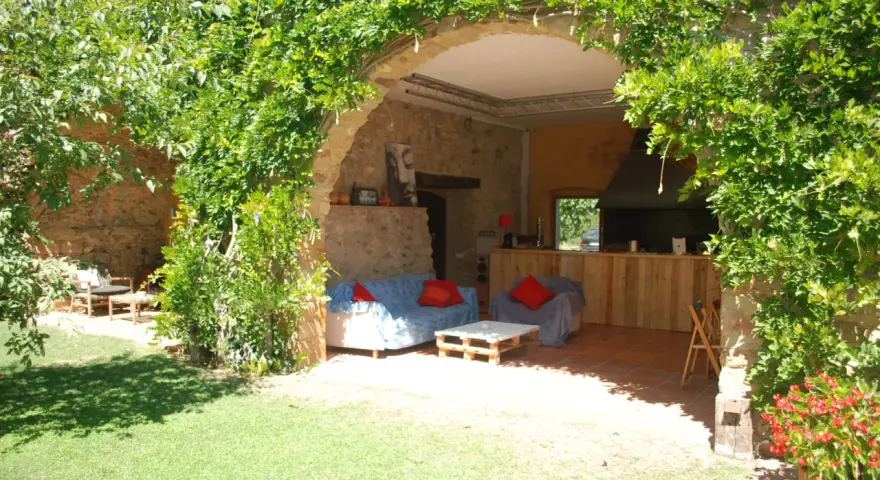 Un jardín con una madreselva planta cubriendo un arco de piedra. Dentro, sofás y una barra de madera.