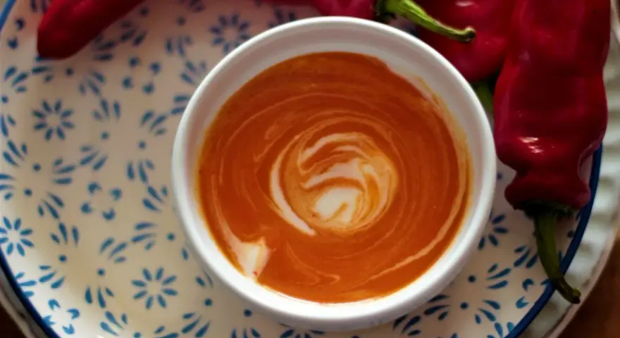 Crema di peperoni vellutata, con un vortice di panna, accanto a peperoni rossi freschi su un piatto decorato.