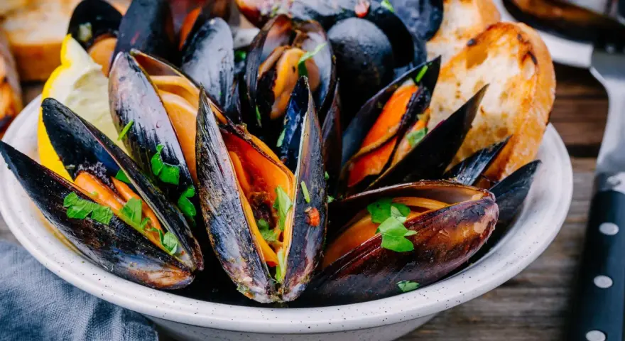 Mejillones en salsa, servidos en un cuenco blanco con perejil fresco, rodajas de limón y pan tostado.