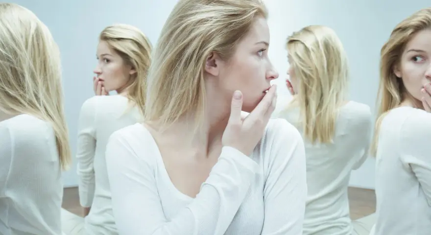 Kobieta z blond włosami w białej bluzce, otoczona swoimi odbiciami w lustrze.