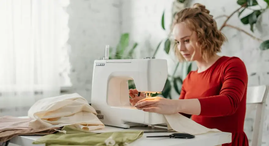 Kobieta w czerwonej sukience tworzy ubrania przy maszynie do szycia. Na stole leżą kolorowe tkaniny.