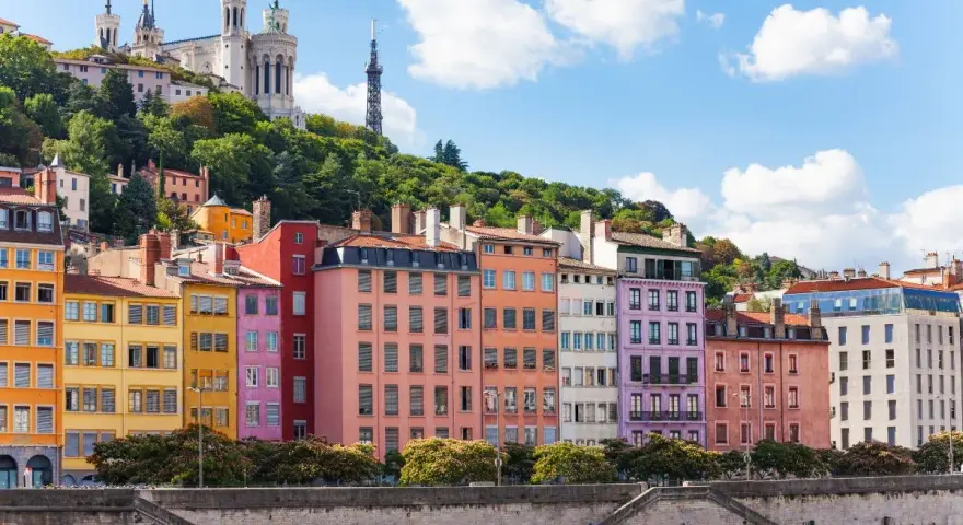Colorful buildings line the river in Lyon, France, with a basilica on the hill above. A perfect scene for Lyon travel.