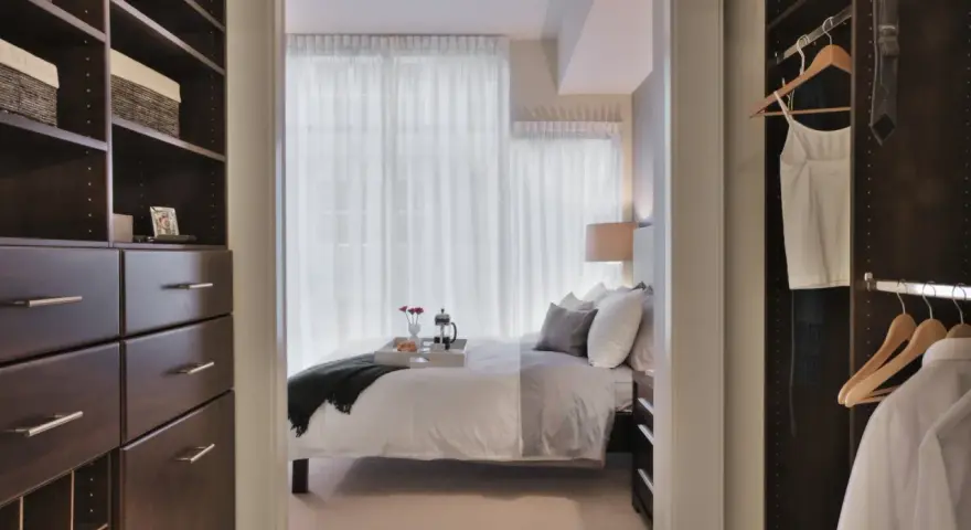 Dormitorio con vestidor integrado. Un espacio organizado con estanterías oscuras y cajones, que da paso a una cama acogedora y un armario con ropa colgada.